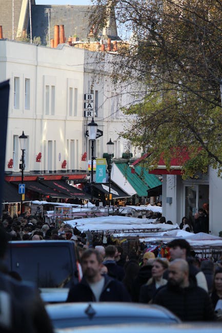 A busy street scene in NW6 near Queens Park Farmers Market showing a row of multi-storey terraced buildings with colorful facades, some with awnings over shopfronts. The street is lined with parked ca